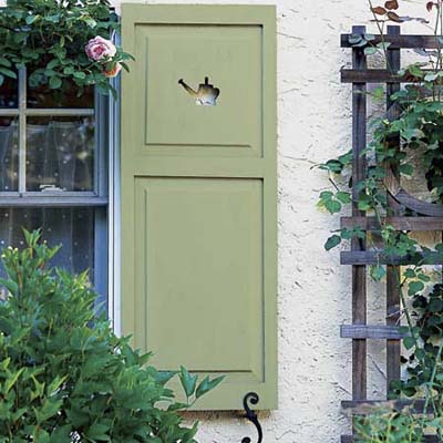 green shutter has a watering can cut out and a S- shaped iron shutter dogs. A trellis is near the shutter