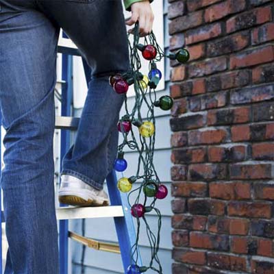 person standing on ladder in front of a house with string of holiday lights in one hand