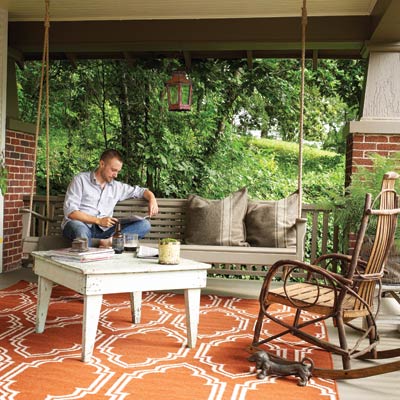 a porch with porch swing, outdoor furniture and area rug, man sits reading on the porch swing