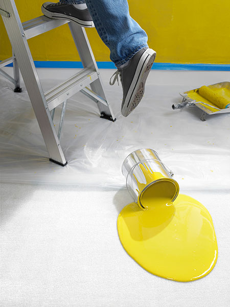 bright yellow paint spilled onto the floor next to a someone's feet stepping off a ladder and a paint roller and pan in the background next to a taped off wall painted the color of the spilled paint