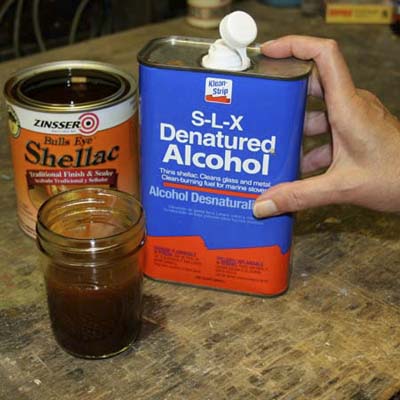 small jar with mixture in front of can of shellac and hand on can of denatured alcohol