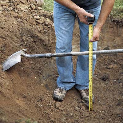 man with tape measure checking the depth of a hole by measuring from the shovel handle laid across the hole